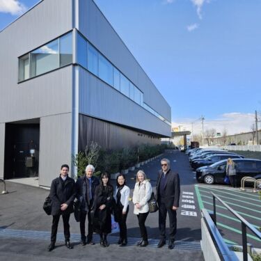 A group of six people, dressed in winter coats and business attire, stand in front of a modern, gray, two-story building with large windows. They are smiling and posing for the photo. The building has a parking lot to the right with several parked cars. The sky is clear and blue.