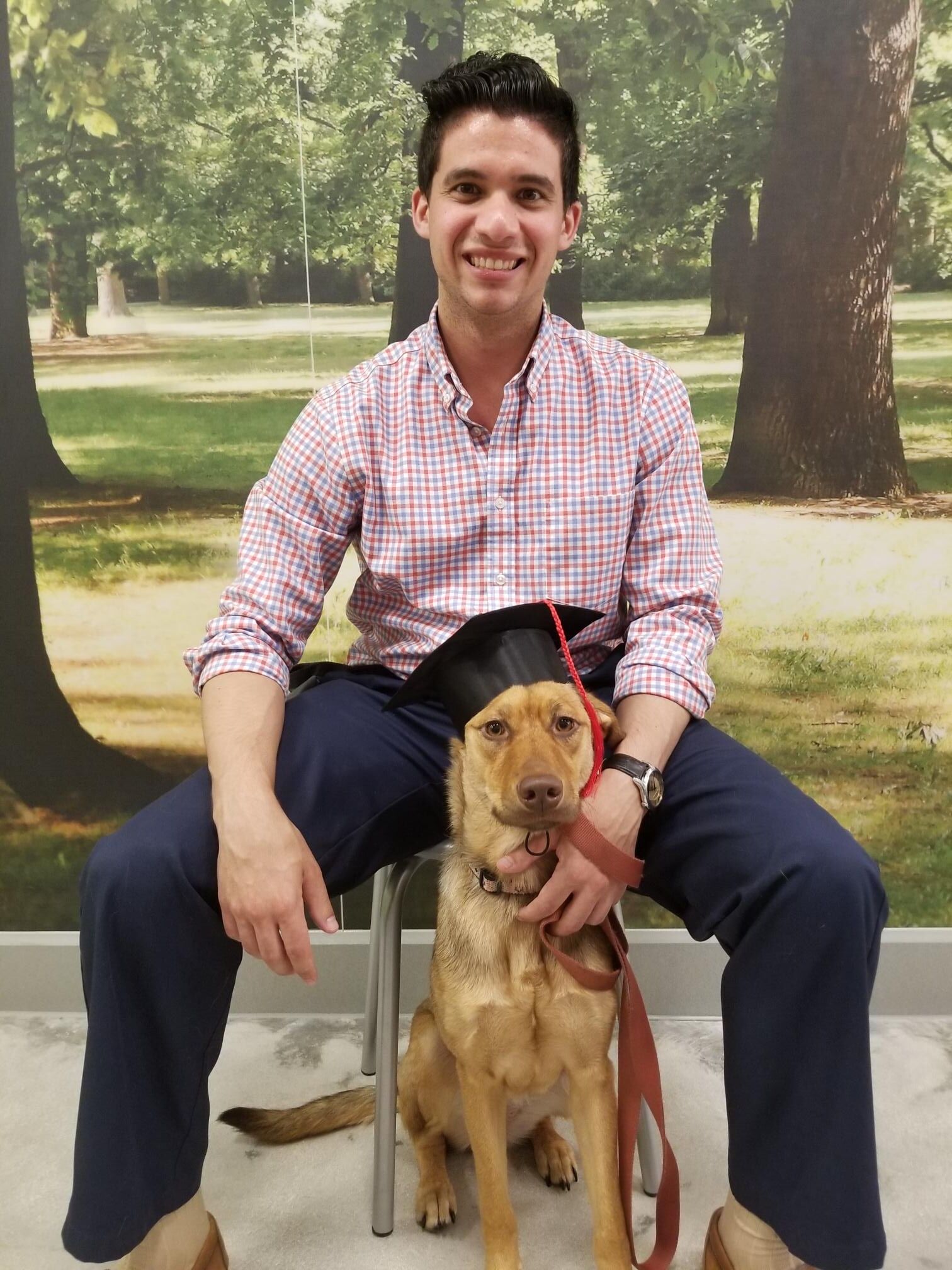 A man in a checkered shirt and navy pants sitting on a stool with a light brown dog sitting between his legs. The dog is wearing a black graduation cap with a red tassel. Both the man and the dog are looking at the camera, with the man smiling.